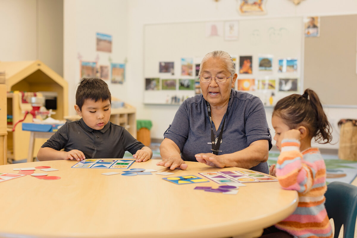 Head start educator Jacqueline Toledo-Magdalena sitting at a classroom table with two children, engaging in an activity together.