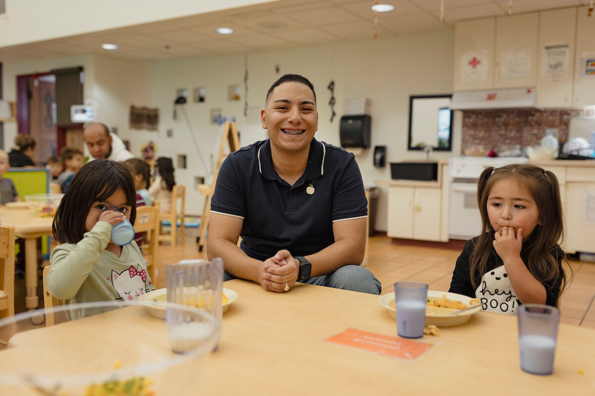 PreK Educator Randy Orona sitting at a lunch table with two children eating their meals.